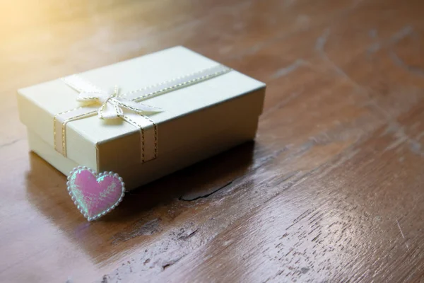 Primer plano pequeña caja de regalo de turba blanca con cinta blanca y el corazón diminuto brillo rosa con la luz naranja en la mesa de madera áspera marrón. Copia sapce para edición y texto . — Foto de Stock