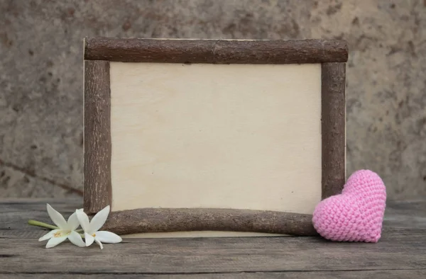 Marco de madera marrón en la mesa de madera áspera con la millingonia blanca y el corazón de punto rosa. Fondo de la pared de roca. La luz del sol brilla en el marco. Copiar espacio para editar y texto . — Foto de Stock