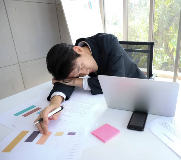 Young handsome Asian business man fall asleep on the working desk because of tried of workload. Silver laptop, mobile phone, pink notepad, and other documents also place on. — Stock Photo, Image