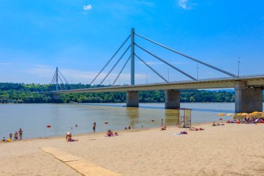 Novi Sad, Sırbistan - 3 Temmuz 2019: İnsanlar Sırbistan 'ın Novi Sad kentindeki Liberty Köprüsü manzaralı Tuna Nehri üzerindeki bir plajın keyfini çıkarıyor
