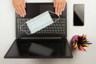 Medikal maske dizüstü bilgisayarın klavyesinde beyaz tahta bir arka planda renkli kalemlerin yanında duruyor.