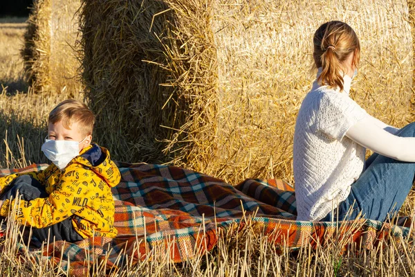 mom and baby sit on the ground in nature on the background of haystacks in medical masks