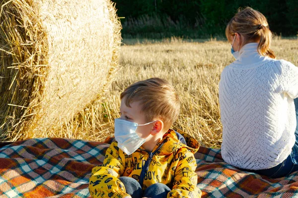mom and baby sit on the ground in nature on the background of haystacks in medical masks