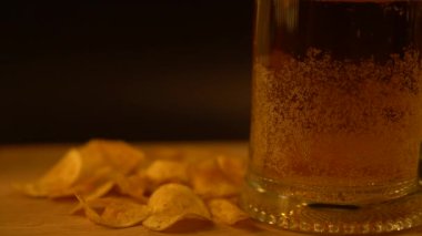 Bardakta bira ve saçılmış cipsler siyah arka planda tahta bir tahtanın üzerinde.