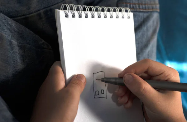 lonely white orphan boy in jeans, faceless, draws a house in a notebook with a liner copyspace
