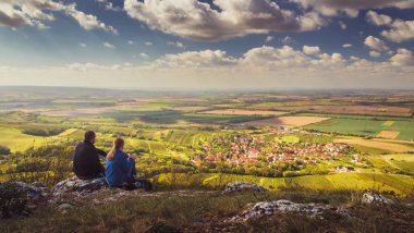 Landscape - Two people (couple - man and woman) sitting on a mountain on a stone and looking into the valley on beautiful autumn landscape with village, vineyards and fields. Blue sky, white clouds. Palava, Moravia, Czech republic clipart