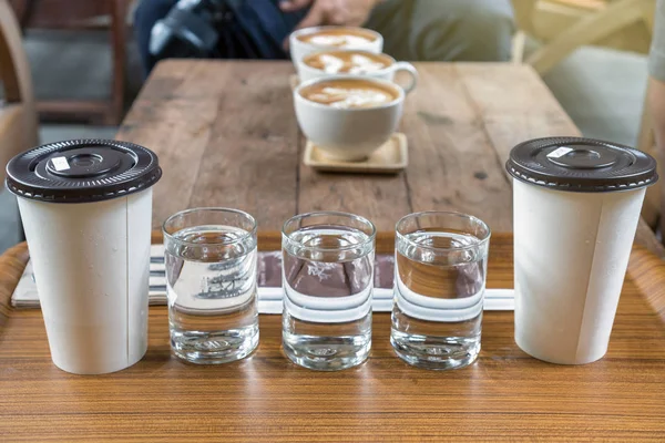 Coffee cup set which consist of glassed of water and ice coffee in paper cup over the photo blurred of latte art hot coffee on the wooden table, drink and coffee shop concept