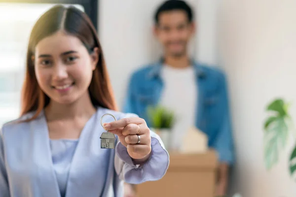 Closeup Asian young woman hand holding the house key chain over the photo blurred of lover carrying big cardboard box for moving in new place after sign contact, Moving and House Hunting concept