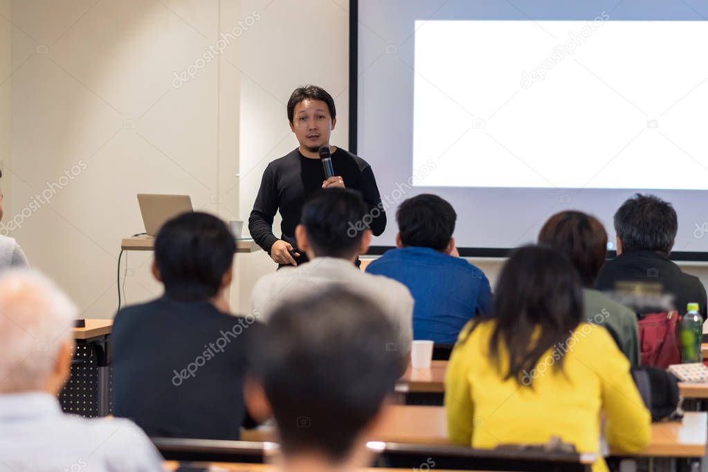 Asian Speaker with casual suit on the stage in low light over the presentation screen in the business or education seminar