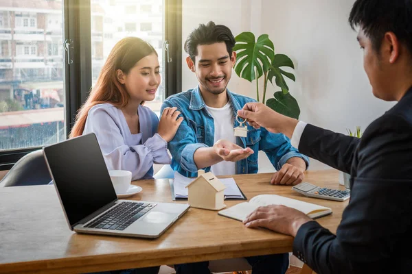 Sale representative offer the house key chain to Asian young couple after signed contact house purchase or rental on the working space table in new house,Moving and House Hunting concept