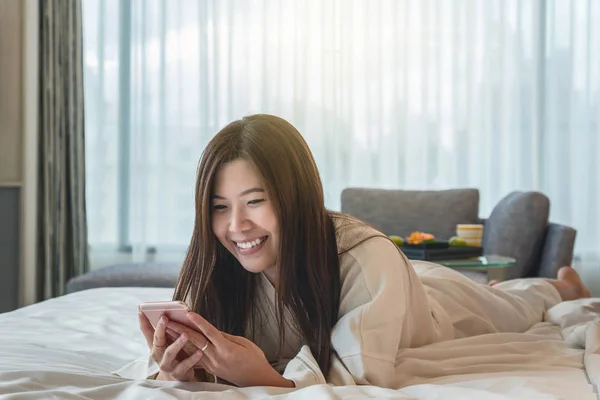 Asian woman in bathrope suit using the smart mobile phone with smiling action on the bed when wake up in luxury hotel, lifestyle and leisure concept