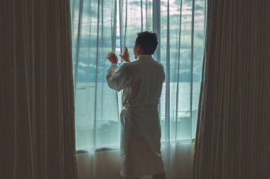 Bornoz giymiş Asyalı bir adam sabah lüks bir otelde yataktan kalkıp tatil konsepti ve yaşam tarzıyla deniz sahilini geziyor.