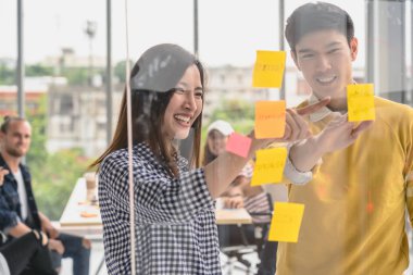 Bir çift yaratıcı iş arkadaşı, modern işyerinde Asyalı İş Adamları Grubu 'nun fikirlerini gündelik kıyafetlerle sunarken, sütçü kurulundaki postit' i işaret ediyorlar.