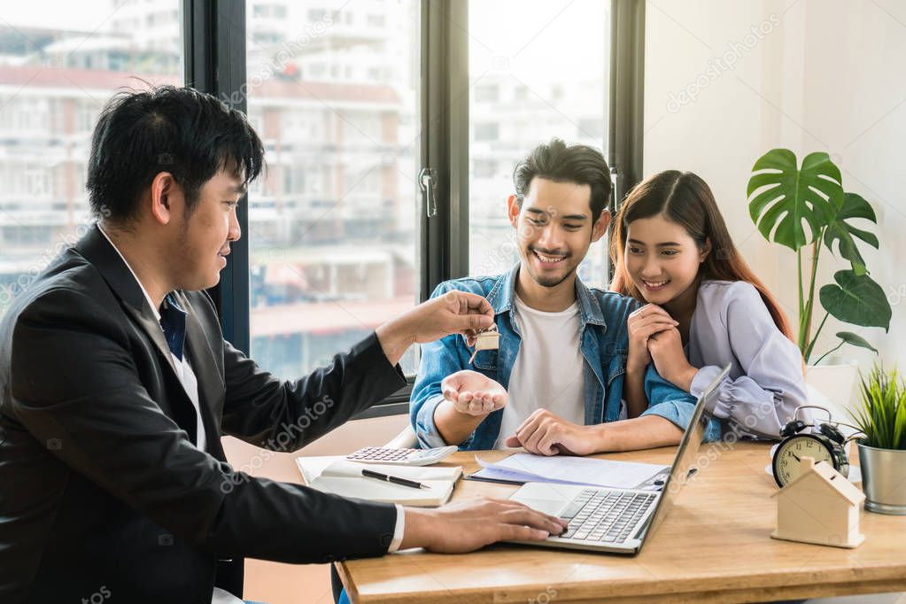 Sale representative offer the house key chain to Asian young couple after signed contact house purchase or rental on the working space table in new house,Moving and House Hunting concept