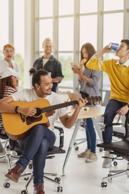 Asian worker playing the guitar over the Group Of Asian and Multiethnic Business people with casual suit talking and eatting with happy action when lunch time in the creative office workplace, life style and relax work concept clipart