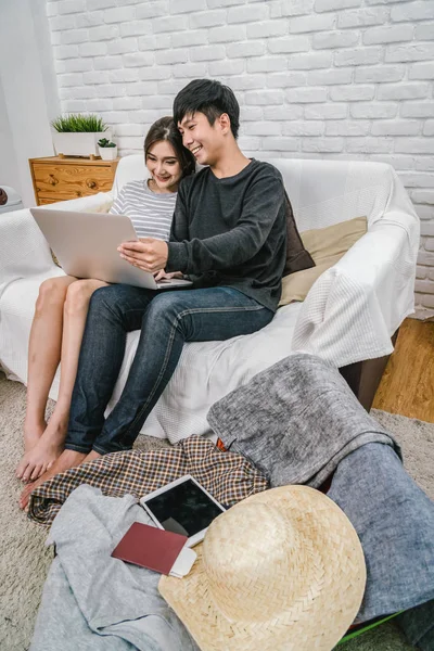 Happy Asian Couple planning and booking the hotel for travel with technology laptop on the sofa of living room at the modern home, Couple, life style and traveler concept,