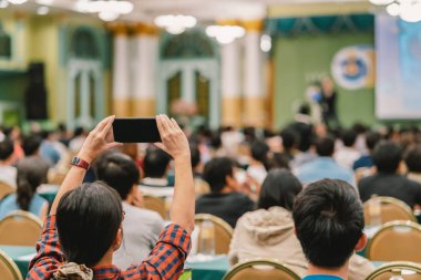 Fotoğraf çekme veya Asya hoparlör için rahat kıyafet ile sahne alanı'nda mevcut konferans salonu veya Seminer Toplantı içinde belgili tanımlık perde akış canlı yapmak için akıllı cep telefonu tutan Closeup kadın izleyici el