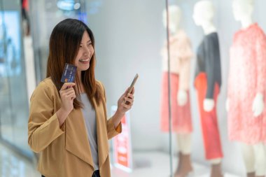 Asyalı bir kadın mağazadan alışveriş yapmak için cep telefonu kullanıyor. Giyim mağazası geçmişi, teknoloji para cüzdanı ve online ödeme konsepti, kredi kartı modeli.