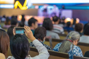 Fotoğraf çekme veya Asya hoparlör için rahat kıyafet ile sahne alanı'nda mevcut konferans salonu veya Seminer Toplantı içinde belgili tanımlık perde akış canlı yapmak için akıllı cep telefonu tutan Closeup kadın izleyici el