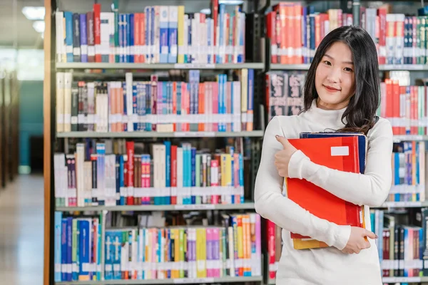 身着休闲套装的亚洲青年学生站在大学图书馆的书架上 拿着书架 有各种书籍背景 回到学校的概念 — 图库照片