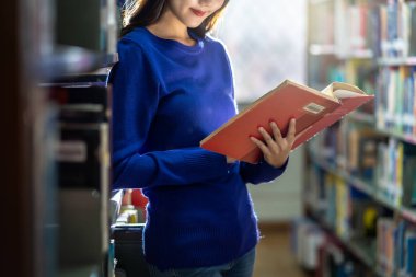 Yakın plan Asyalı genç öğrenci ayakta duruyor ve el ele kitabı tutuyor ve kütüphanede kitap rafında okuyor çeşitli kitap geçmişi olan, okul konseptine geri dönüyor.