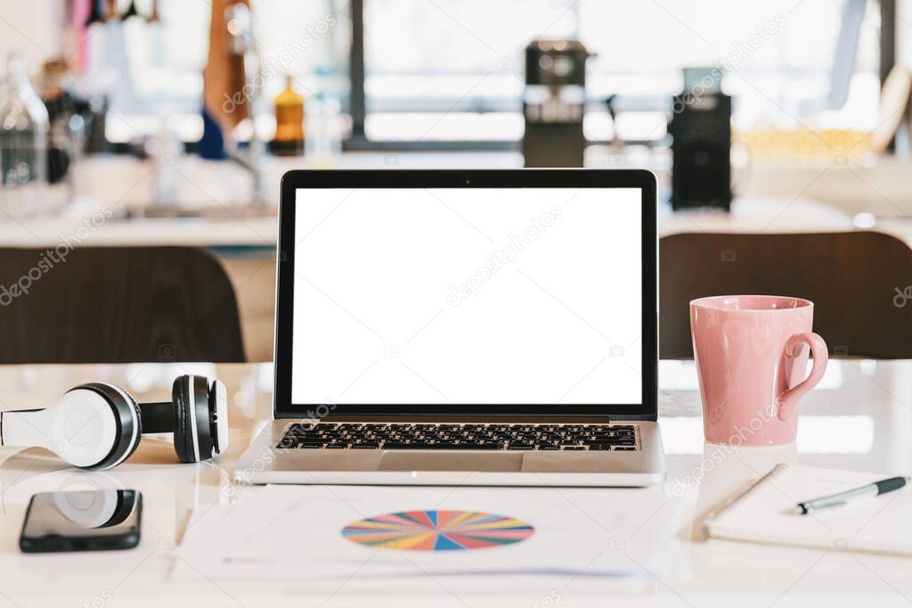 Blank white screen of laptop with headphone, coffee cup, mobile phone and data graph document on the table in kitchen of modern house, business and creative design concept, device for video conferenc