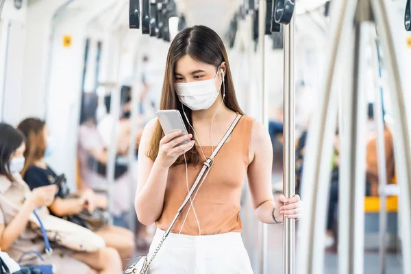 Covid19大都市での旅行時に地下鉄の電車の中でスマート携帯電話を介して外科マスクとリスニング音楽を身に着けている若いアジアの女性の乗客発生 感染症と流行の概念 — ストック写真