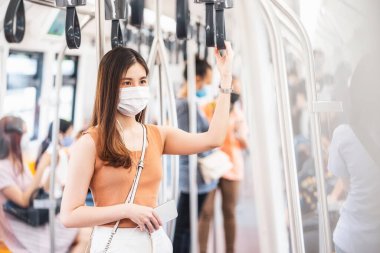 Young Asian woman passenger wearing surgical mask and listening music via smart mobile phone in subway train when traveling in big city at Covid19 outbreak, Infection and Pandemic concep clipart