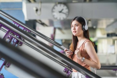 Genç Asyalı kadın yolcu kulaklık takıyor ve müzik dinlemek için akıllı cep telefonu kullanıyor ve büyük şehirde seyahat ederken metro istasyonunda merdivenlerden yukarı çıkıyor, banliyö ve ulaşım konsepti