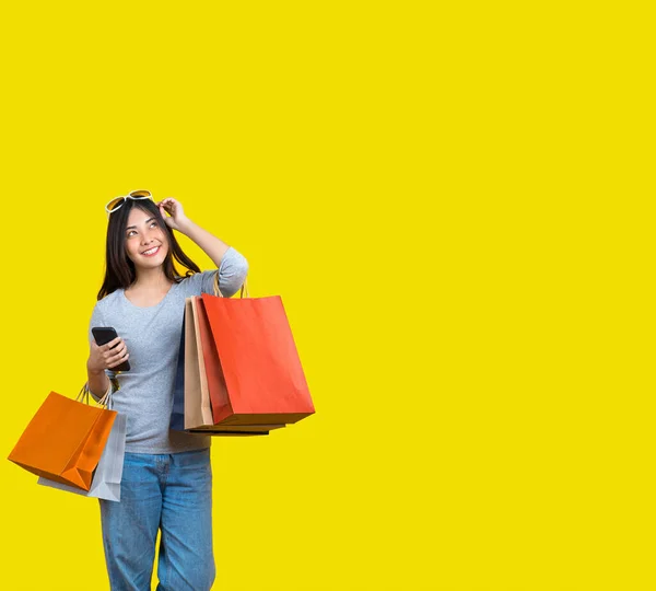 Banner of Attractive Asian smiling young woman Carrying a shopping coloful bag, mobile phone and sunglasses on aisolated yellow color background, copy space and studio,black friday season sale concept