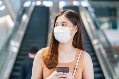 Genç Asyalı kadın yolcu, cep telefonlu cerrahi maske takıyor.
