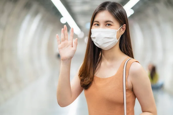 若いですアジアの女性旅客身に着けています外科マスクと挨拶にカメラで彼女の友人で地下鉄インターチェンジで大都市での旅行時Covi19発生 新しい通常の概念 — ストック写真
