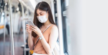 Genç Asyalı kadın yolcu cerrahi maske takıyor ve metroda akıllı cep telefonuyla müzik dinliyor Covid19 salgını, enfeksiyon ve salgın konsepti ile büyük bir şehirde seyahat ederken
