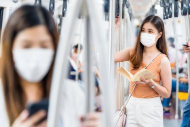 Cerrahi maske takan ve metroda kitap okuyan genç Asyalı kadın yolcu Covid19 salgını, enfeksiyon ve salgın konseptinde seyahat ederken...