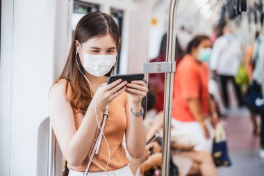 Genç Asyalı kadın yolcu cerrahi maske takıyor ve metroda akıllı cep telefonuyla müzik dinliyor Covid19 salgını, enfeksiyon ve salgın konsepti ile büyük bir şehirde seyahat ederken
