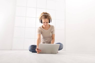 Genç ve kadın ile bilgisayar kulaklıklar, bak beyaz oda pencere arka plan üzerinde izole katta oturan laptop monitör