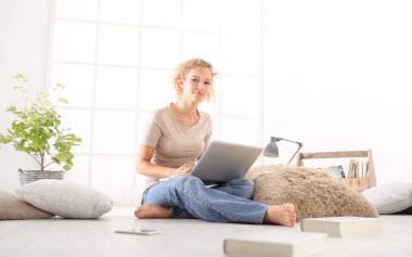 bilgisayar, akıllı telefon ve Kitaplar, arka planda beyaz geniş pencere oturma odasında katta oturan genç kadınla gülümseyen