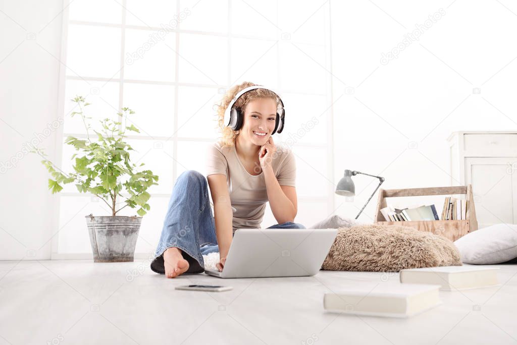 young woman smiling with computer, headphones, smartphone and books, sitting on the floor in living room on white wide window in the background