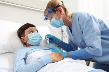 Termometresi olan hemşire hasta yatağında koruyucu güneşlik ve cerrahi maske takan hasta çocuğa ateş ölçer. Corona virüsü Covid 19 koruma kavramı., 