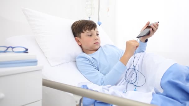 携帯電話のある病院の子供は一人でベッドに横たわっている音楽を聴くためにイヤホンをつけ — ストック動画