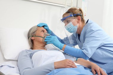 Hemşire, hastane yatağında yatan yaşlı bir kadına koruyucu eldivenler ve maske takarak oksijen maskesi takıyor. Coronavirus Covid 19 koruma kavramı.