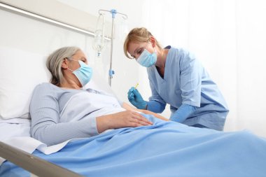 Şırıngalı hemşire aşıyı hastane yatağında yatan yaşlı kadına enjekte ediyor. Koruyucu eldivenler ve tıbbi maske takıyor. Coronavirus covid 19 koruma kavramı.