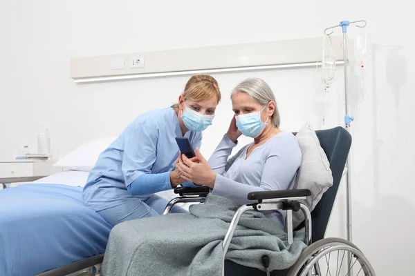 nurse helps with cell phone to contact the elderly lady\'s family in the wheelchair, wearing surgical protective medical masks in hospital room, concept of isolation from corona virus covid 19