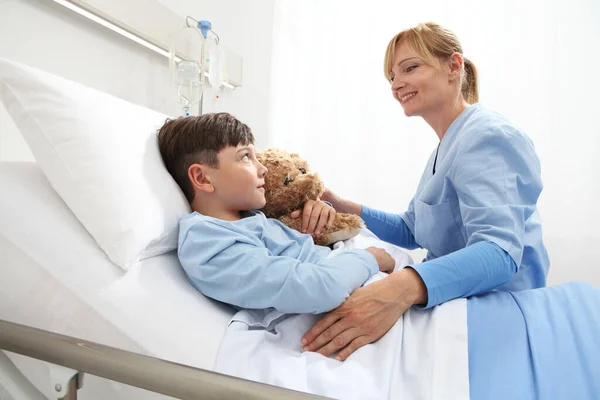 Happy Child lying in bed in hospital room hugging a teddy bear looking nurse smiling and sitting beside him