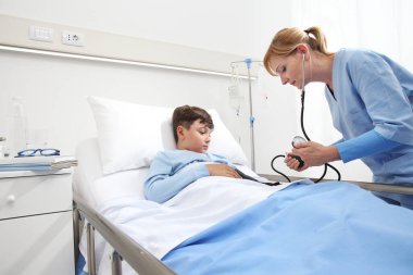 Çocuk hastane odasında yatakta yatıyor ve hemşire kan basıncını sphygmomanometre ve stetoskopla ölçüyor.