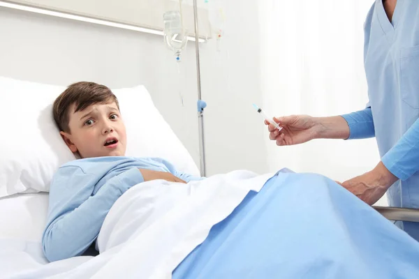 Afraid child lying in bed in hospital room looking at camera scared by the needle of the syringe and nurse giving him an injection