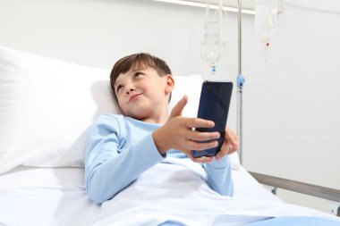 Hastane odasında tek başına yatmış akıllı telefona bakan bir çocuğun portresi. 