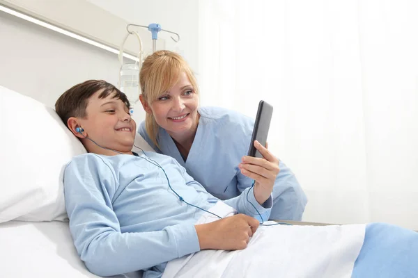 Happy child lying in bed in hospital room and smiling nurse using smartphone surfs the internet wearing earphones