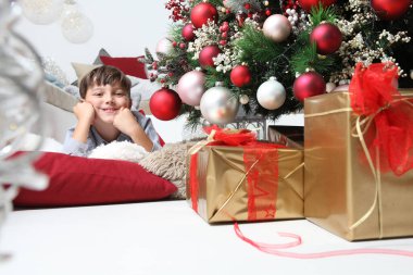 Çocuk ve toplarla süslenmiş Noel ağacı, oturma odasında yerde, paketlenmiş hediyelerin yanında yatıyor.