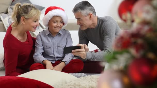 陽気なクリスマス 家で幸せな家族 両親は照明と装飾された木の近くに息子と一緒に携帯電話を見て 喜びと笑顔でリビングルームに座って暖炉で快適 — ストック動画